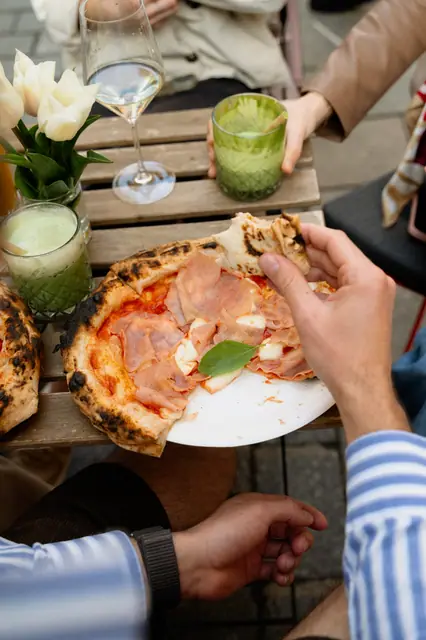 Ruka sahá po kousku pizzy s prosciutto u stolku s drinky