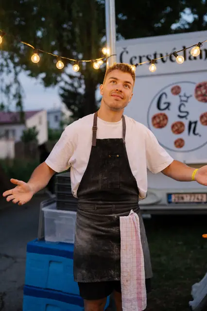 Pizzaiolo s rozpaženýma rukama pod světýlky vedle food trucku s langoši
