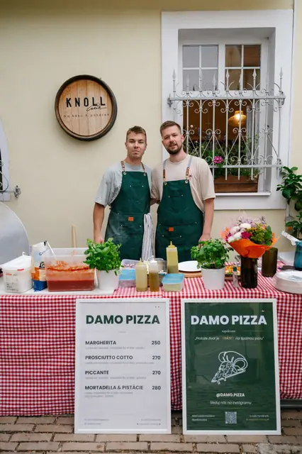Dva pizzaioli DAMO v zelených zástěrách u stánku s kostkovaným ubrusem před kavárnou Knoll
